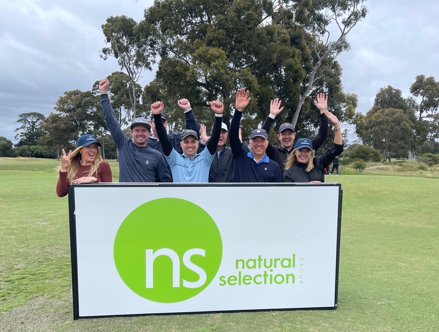 A group of people standing in a line outdoors, smiling and cheering behind a sign that reads