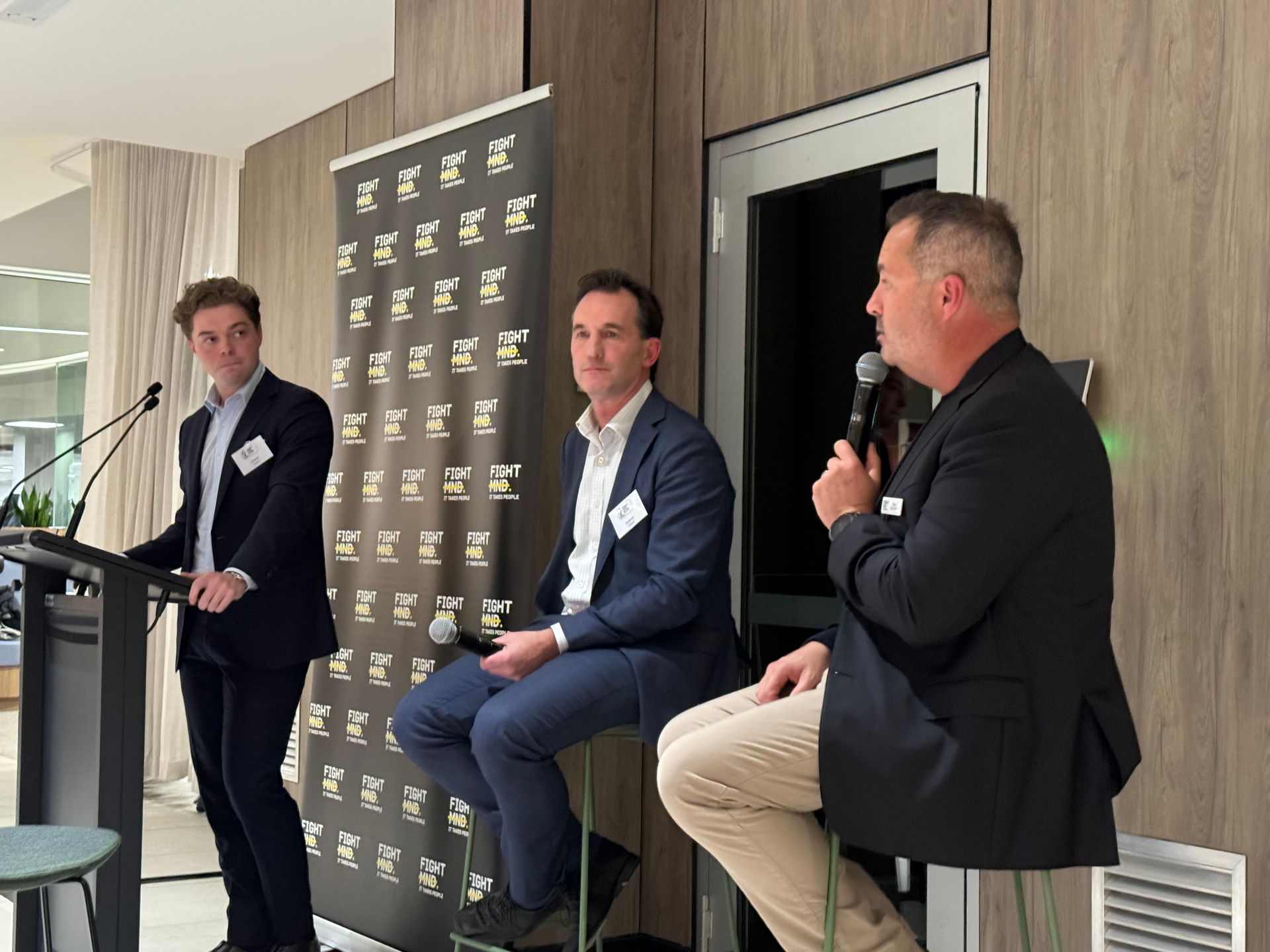 Three professionals in business attire at an indoor panel discussion; one stands at a podium, two sit on stools.