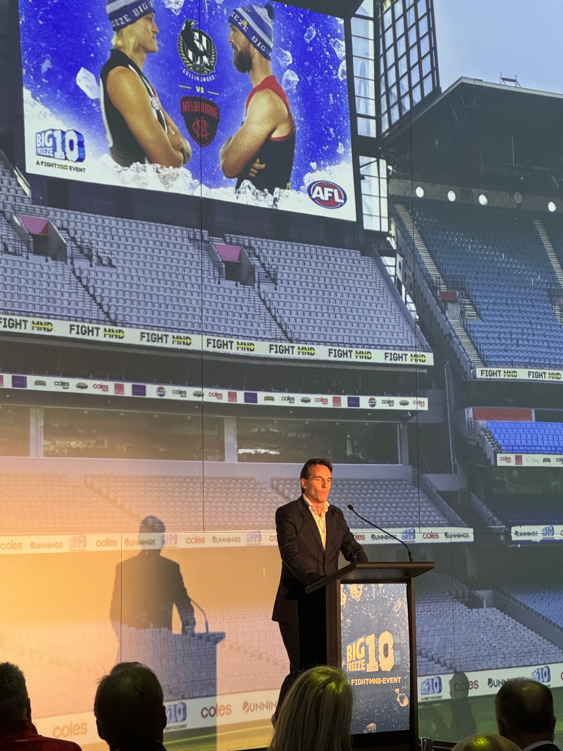 A man speaks at a podium in front of a large screen showing an AFL match graphic inside a stadium.