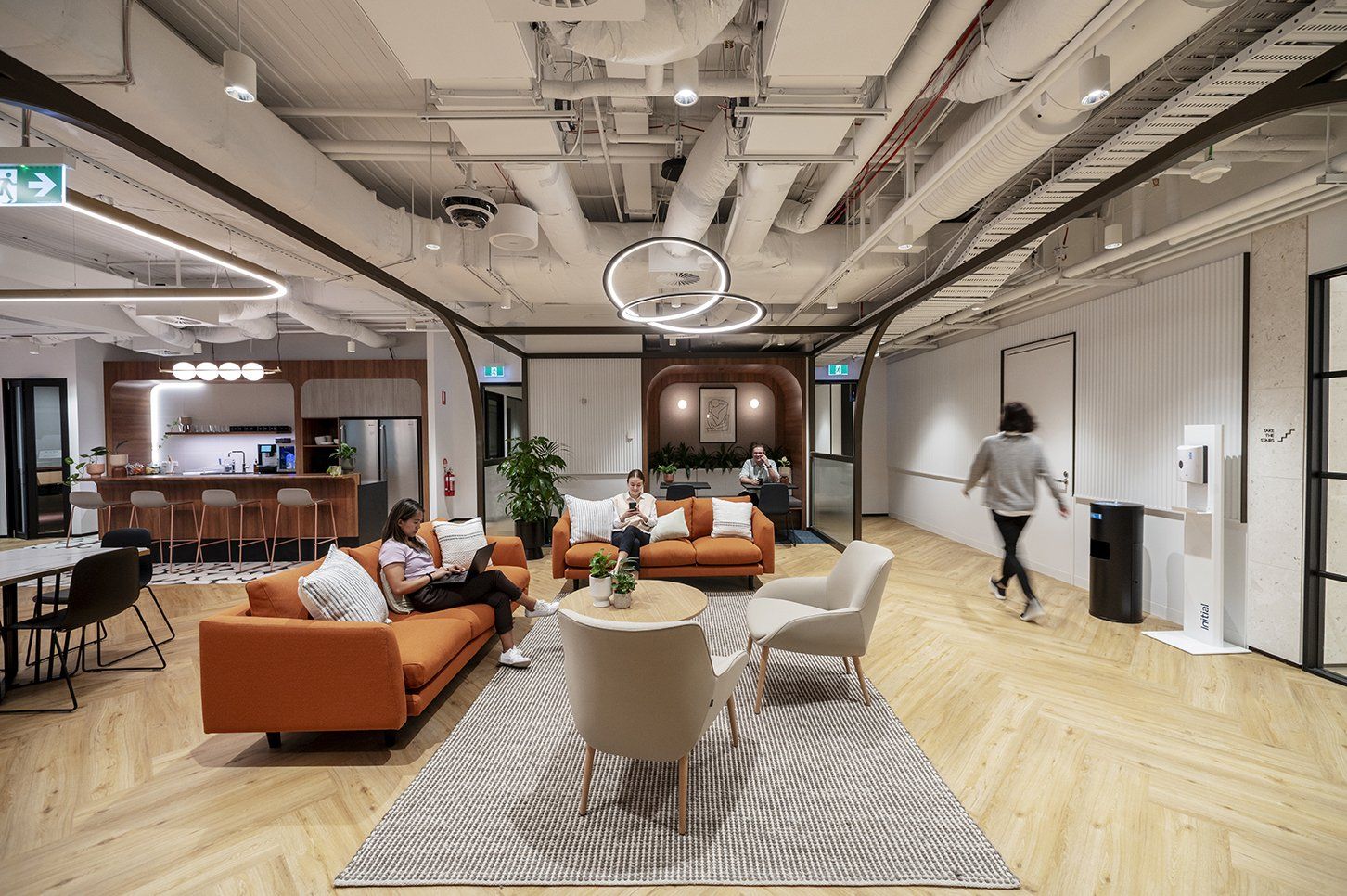 A bright office lounge area with orange sofas, a patterned rug, a kitchen counter, and people working and walking.