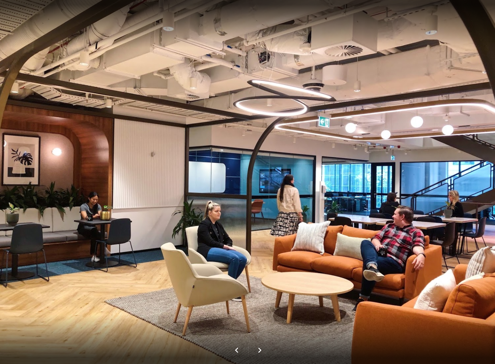A modern, open-concept office lounge with brown sofas, cream armchairs, a coffee table, and people seated in work areas.