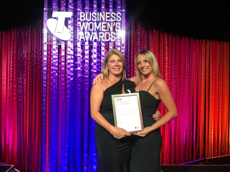 Two people in black evening wear stand smiling on stage, holding an award in front of a purple Telstra Business Awards backdrop.