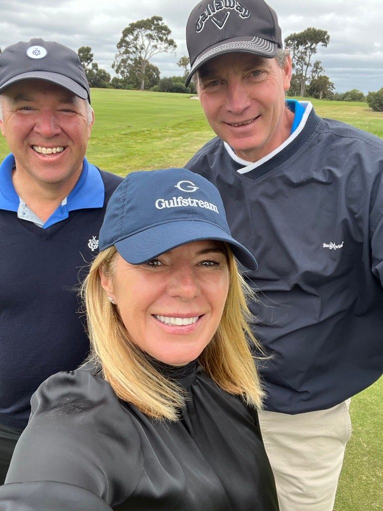 Three people wearing hats smile while posing for a selfie on a sunny, grassy golf course.