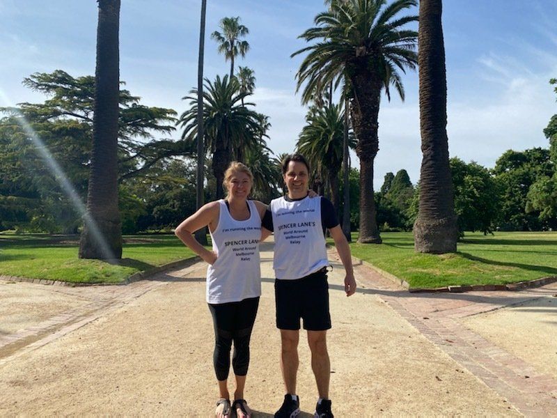 Two people in white athletic tops stand on a gravel path lined with tall palm trees in a park on a sunny day.