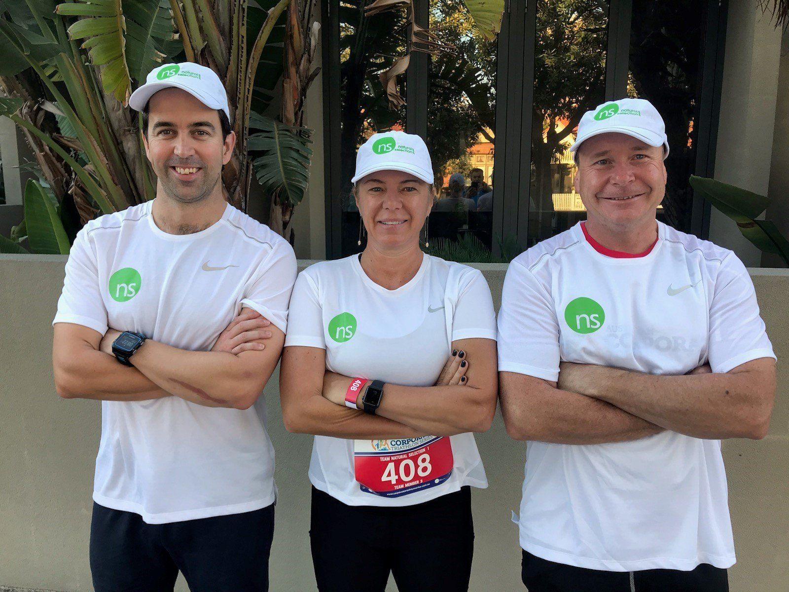 Three people in matching white shirts and caps with green logos stand with arms crossed outdoors in front of a building.
