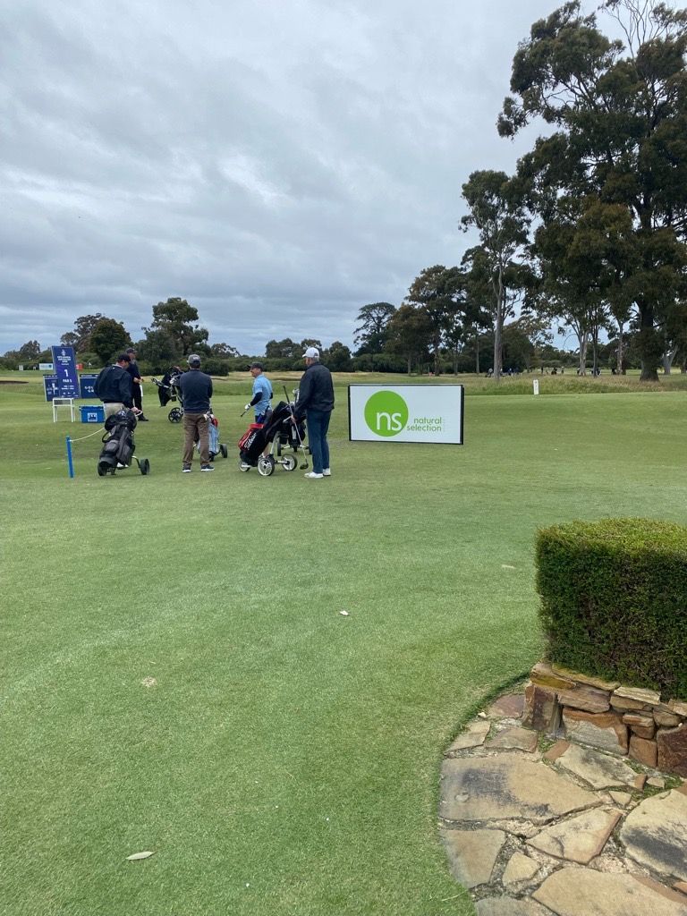 Golfers stand on a green near a sign featuring a circular green logo, with trees and a cloudy sky in the background.