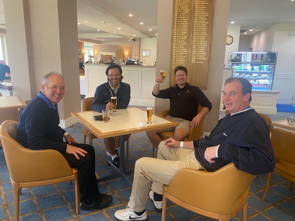 Four people sit at a table in a brightly lit lounge, smiling while drinking beer with a wall plaque visible nearby.