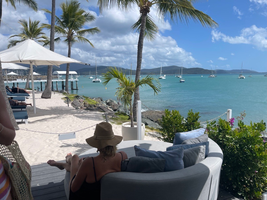 A person in a sun hat relaxes on a lounge sofa overlooking a tropical beach, white umbrellas, and boats in the water.