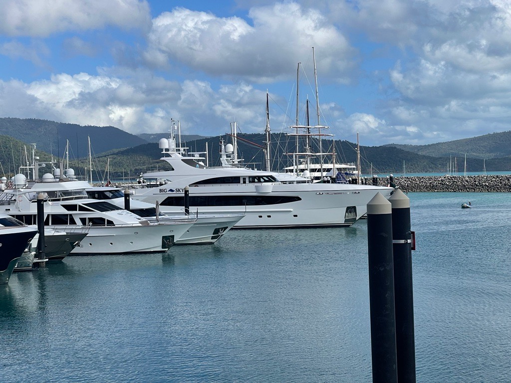 White luxury yachts are moored at a marina with mountains in the background under a cloudy blue sky.