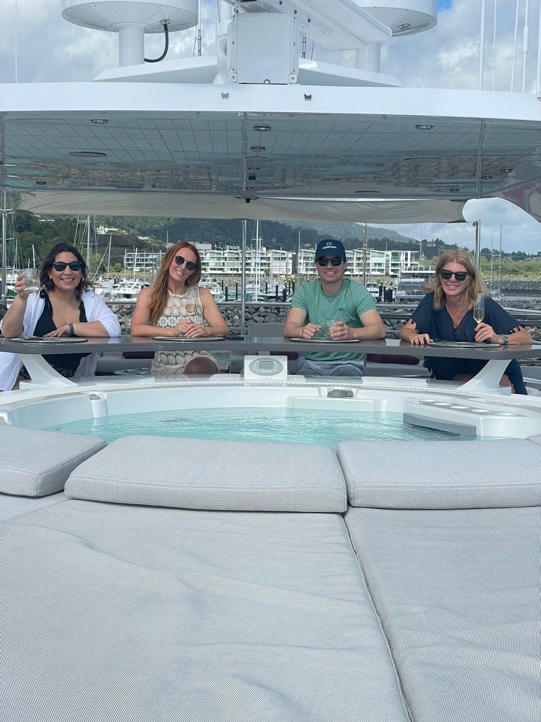 Four people sit at a deck bar on a boat in front of a hot tub, with a marina and hills in the background.