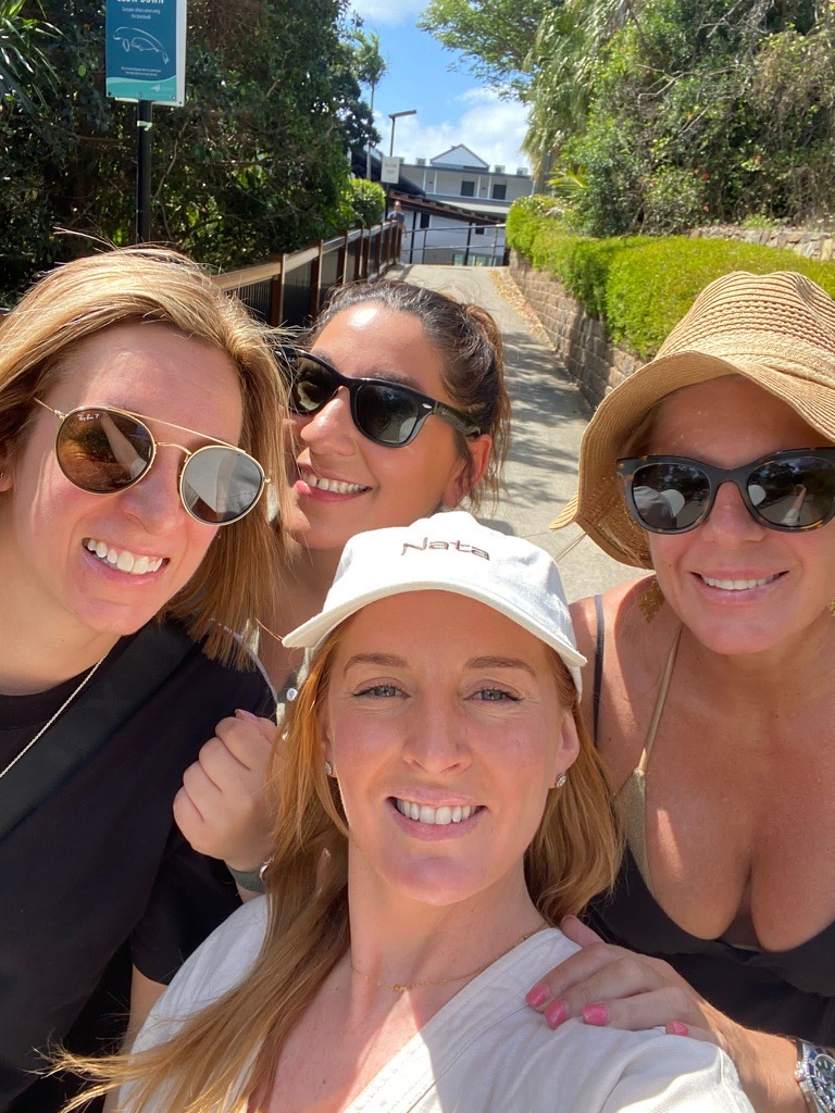 Four people pose for a selfie on a sunny, sloping street lined with green trees and a fence.