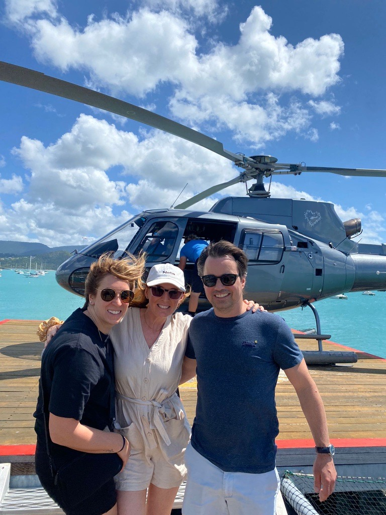 Three people pose for a photo in front of a helicopter on a wooden deck next to turquoise water under a blue sky.