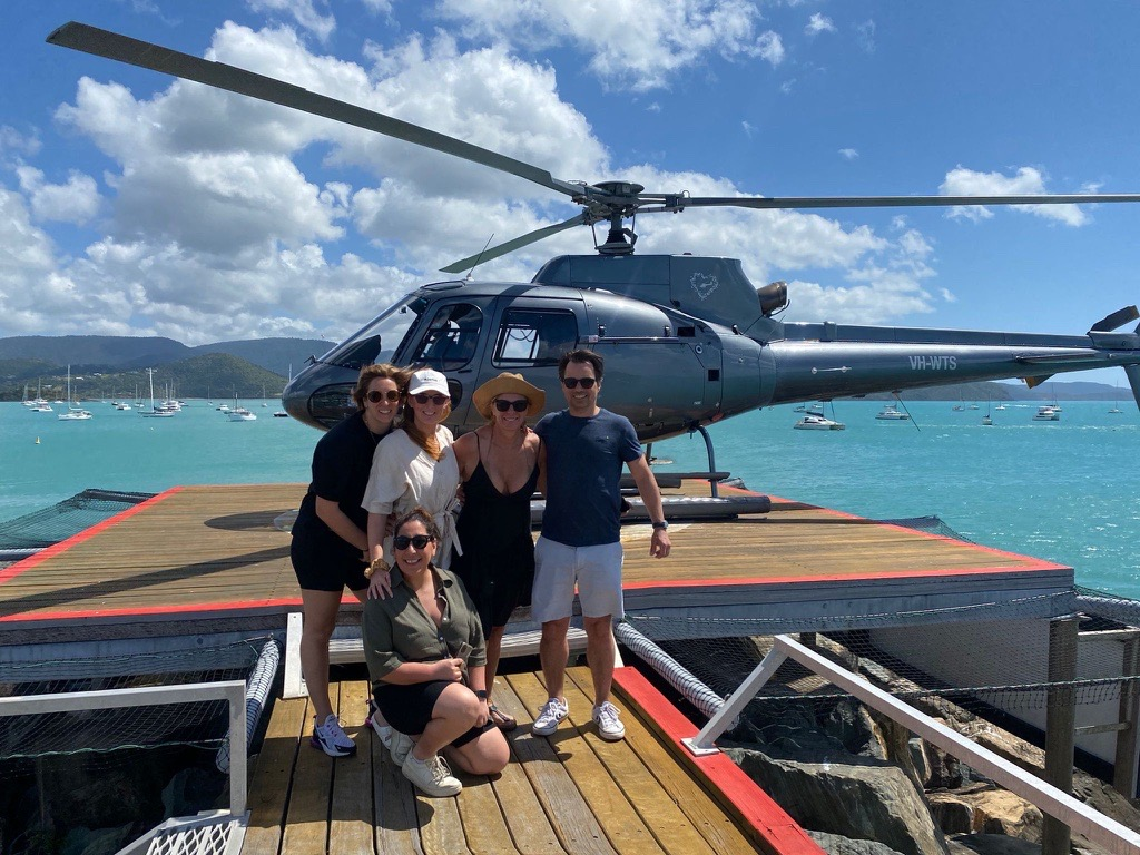 Five people pose for a photo on a wooden helipad next to a helicopter, with a blue ocean and distant coastline behind.