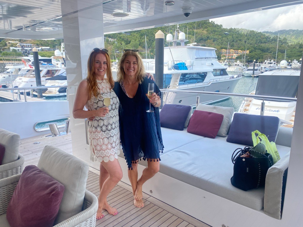 Two smiling people stand on a yacht deck holding glasses, with a marina and boats in the background.