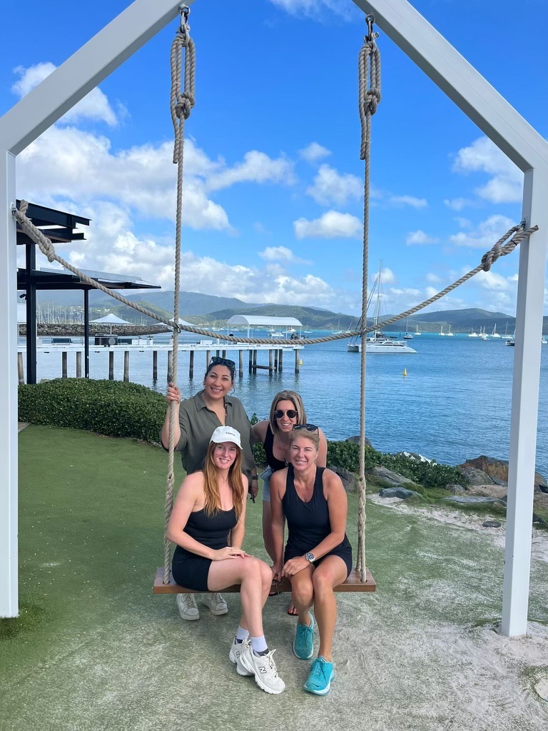Four people pose on a wooden swing frame overlooking a sunny ocean bay with a pier and boats in the background.