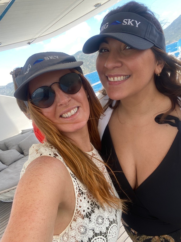 Two women wearing sun visors and smiling while taking a selfie together outdoors on a deck.