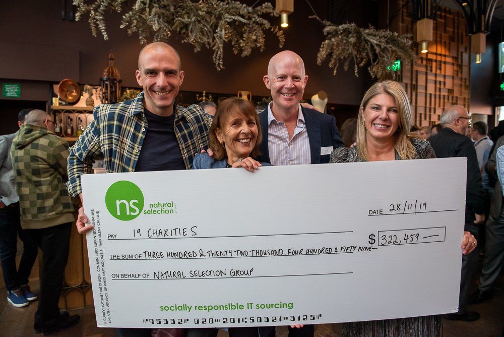 Four people stand smiling while holding a large, ceremonial check from Natural Selection for over $200,000 to 19 charities.