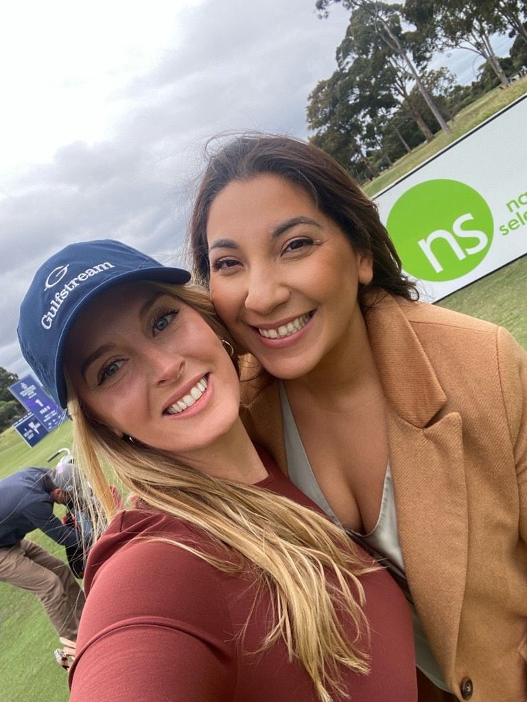 Two smiling individuals take a selfie outdoors at a golf course, with a green and white sponsor sign in the background.