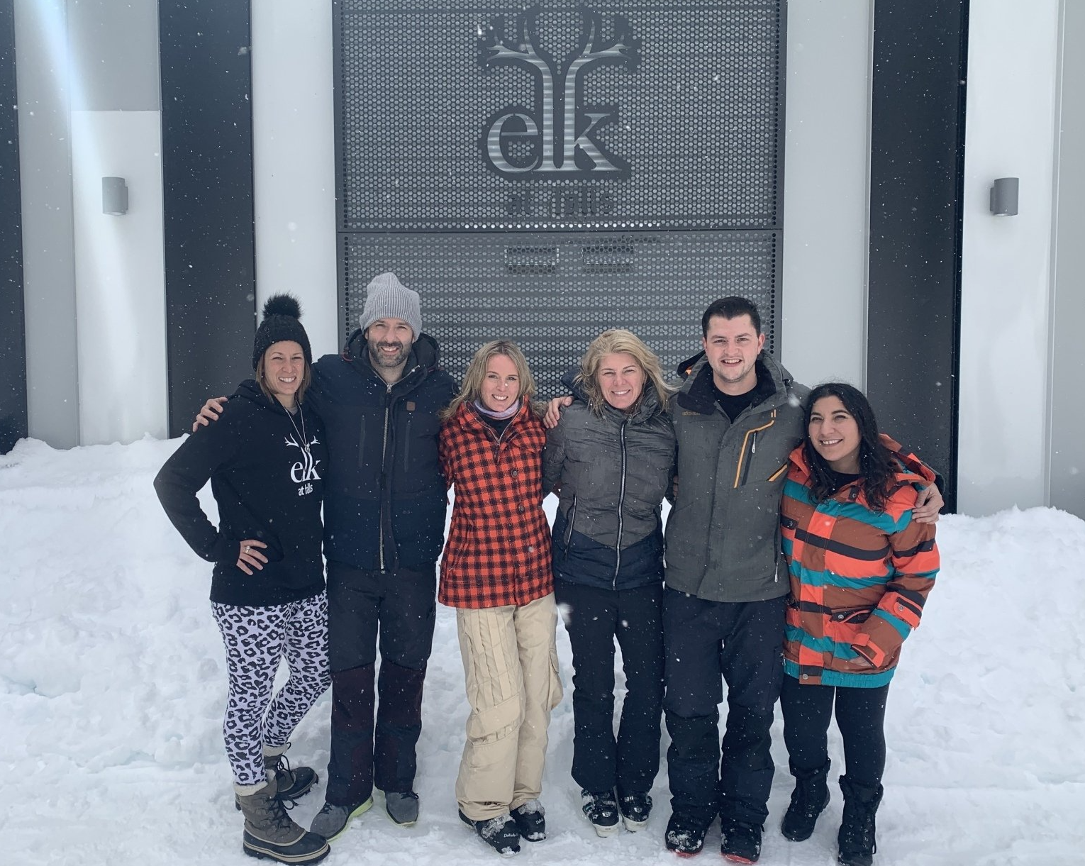 Six people smiling together in front of a snowy building with a metal logo sign reading 