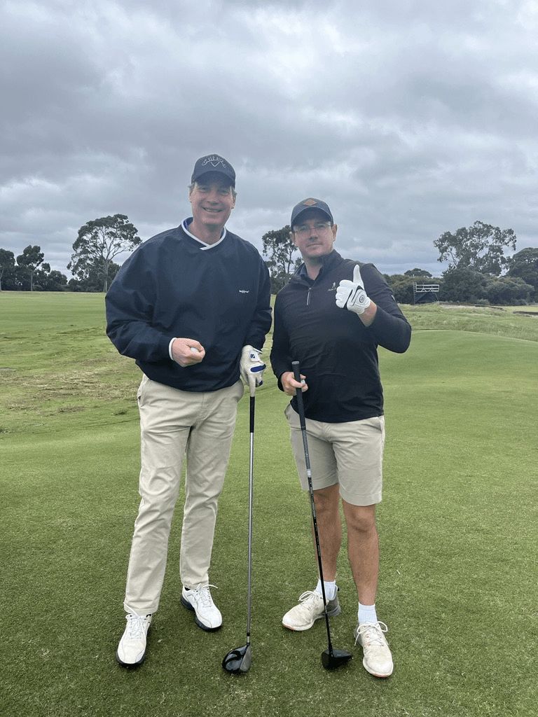 Two golfers in dark jackets and hats stand on a green course, smiling and holding golf clubs.
