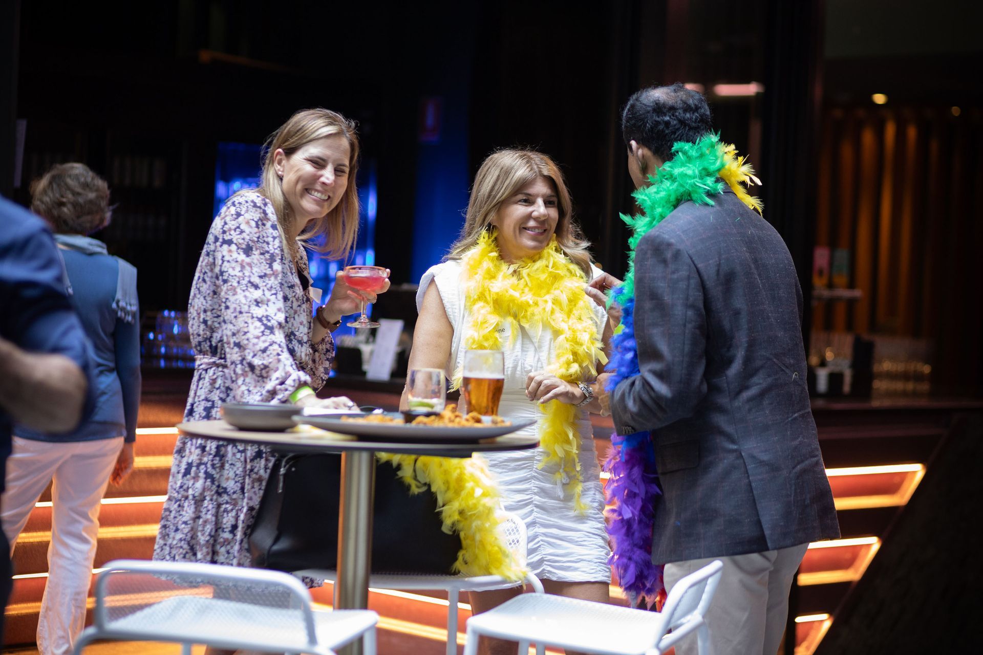 People socialize at a table in a dimly lit bar, wearing colorful feather boas and holding drinks.