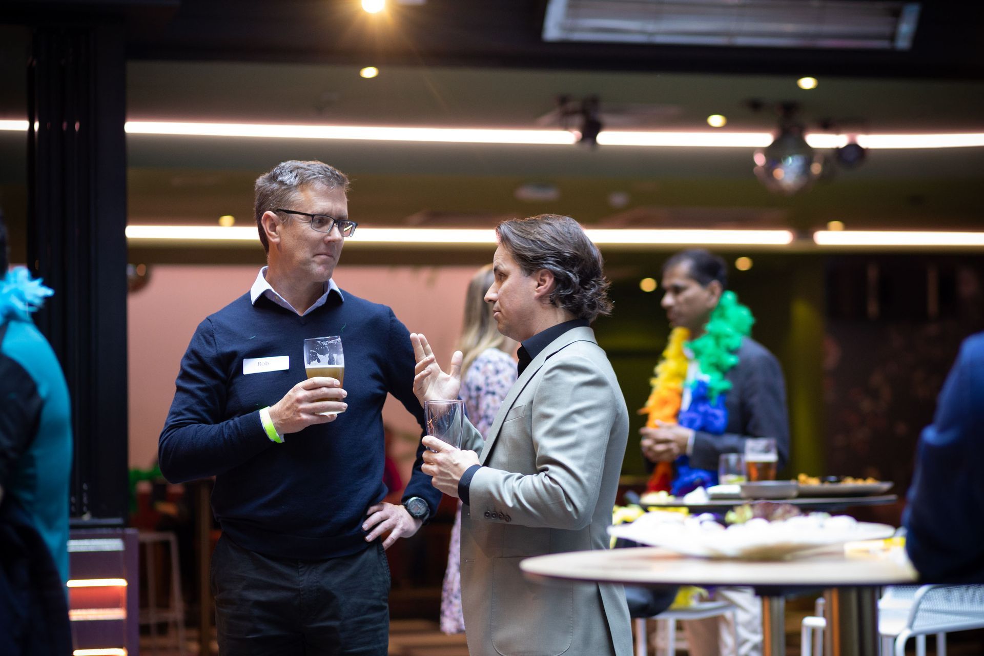 Two people in business attire converse at an indoor social event, with others in the background.
