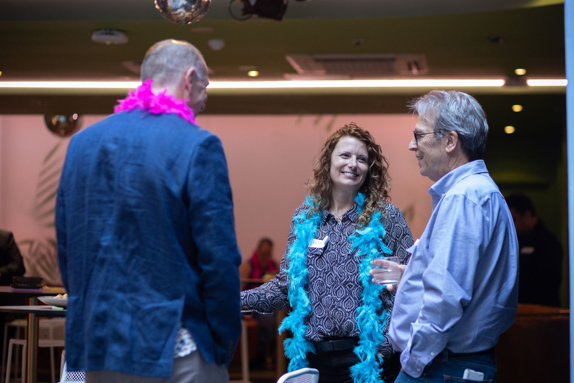 Three people in a social setting; one wears a pink feather boa, another wears a blue feather boa, all are chatting.