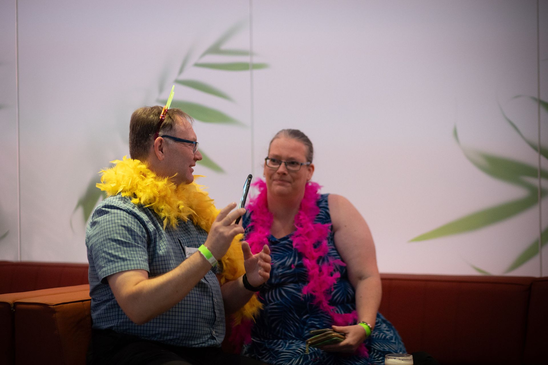 Two people sit on a red couch wearing brightly colored feather boas and talking, against a white wall with leaf decals.