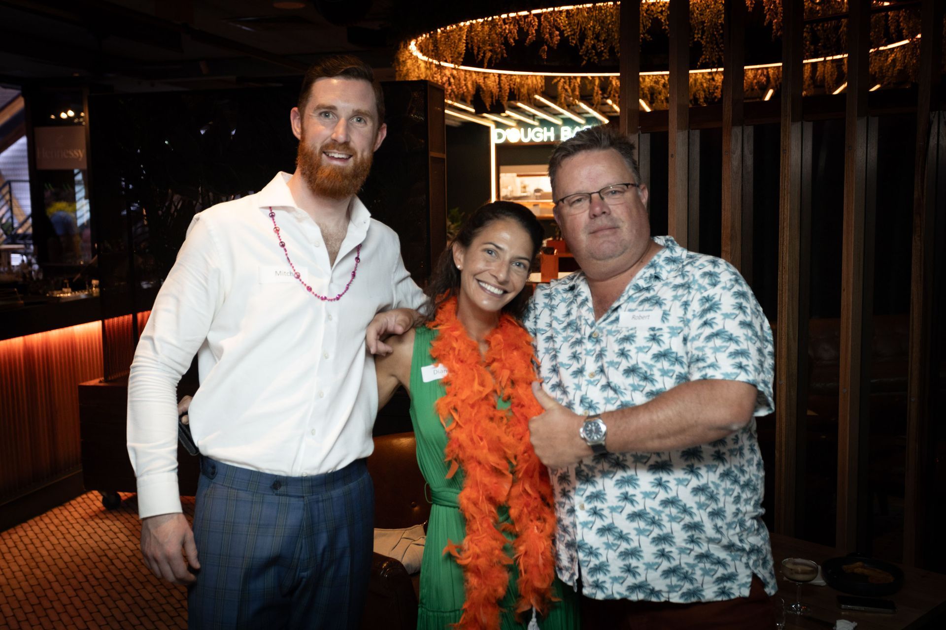 Three people stand in a dimly lit venue, smiling for a photo while wearing colorful leis.