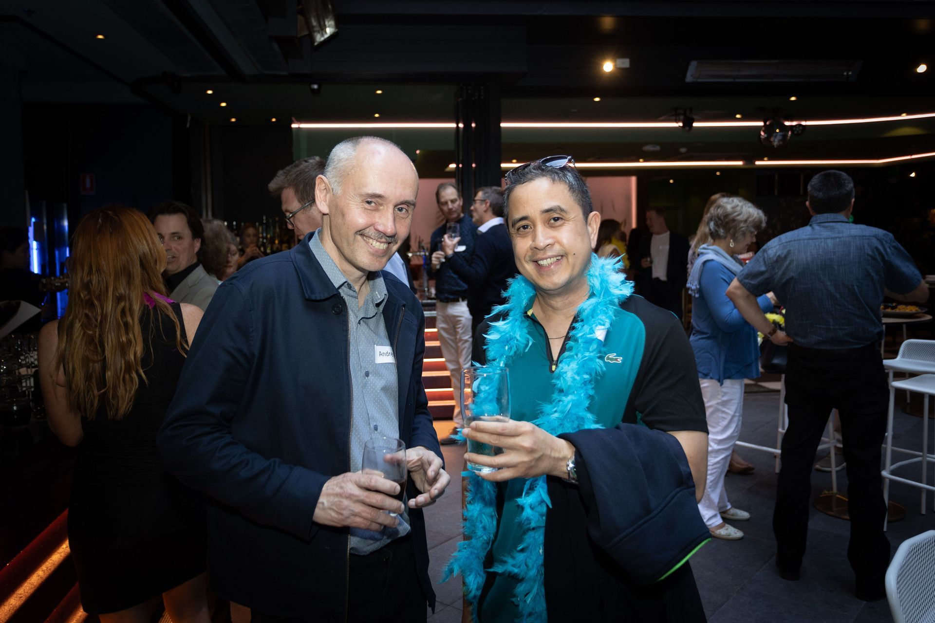 Two men smile while holding drinks at a social event, one wearing a bright teal feather boa; others mingle in the background.
