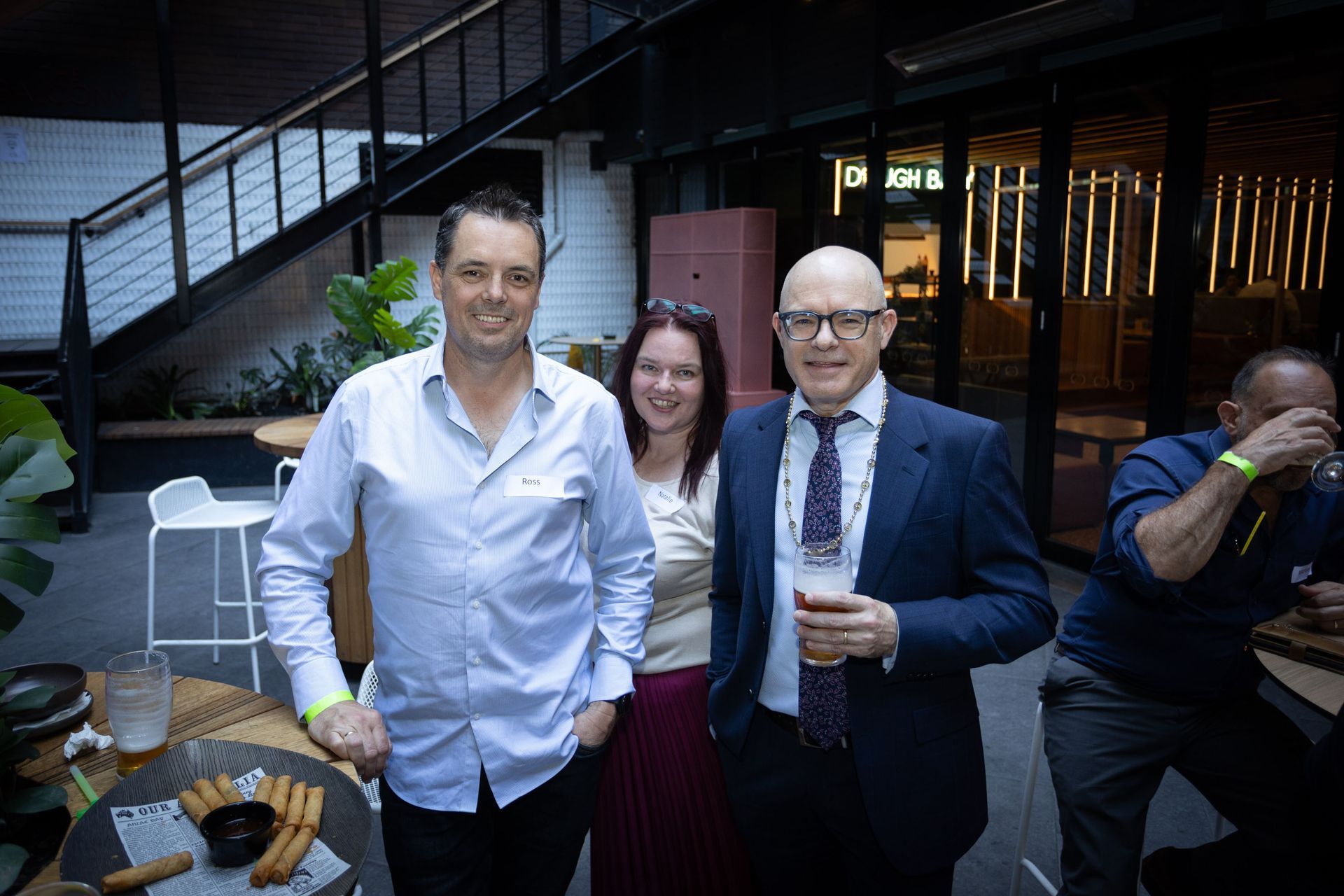Three people socialize at an indoor gathering, with a man in a suit holding a glass in the foreground.