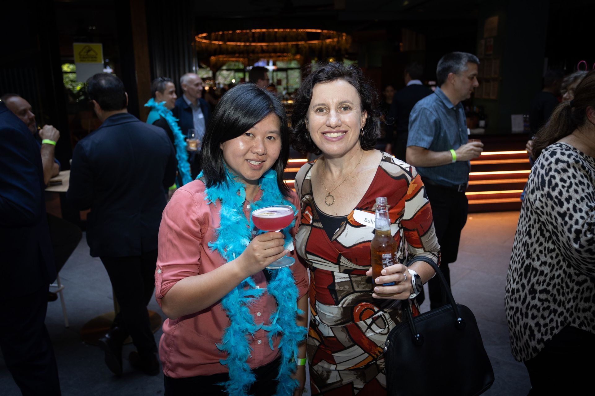 Two smiling women with drinks stand at a party, one wearing a turquoise boa, with guests in the background.