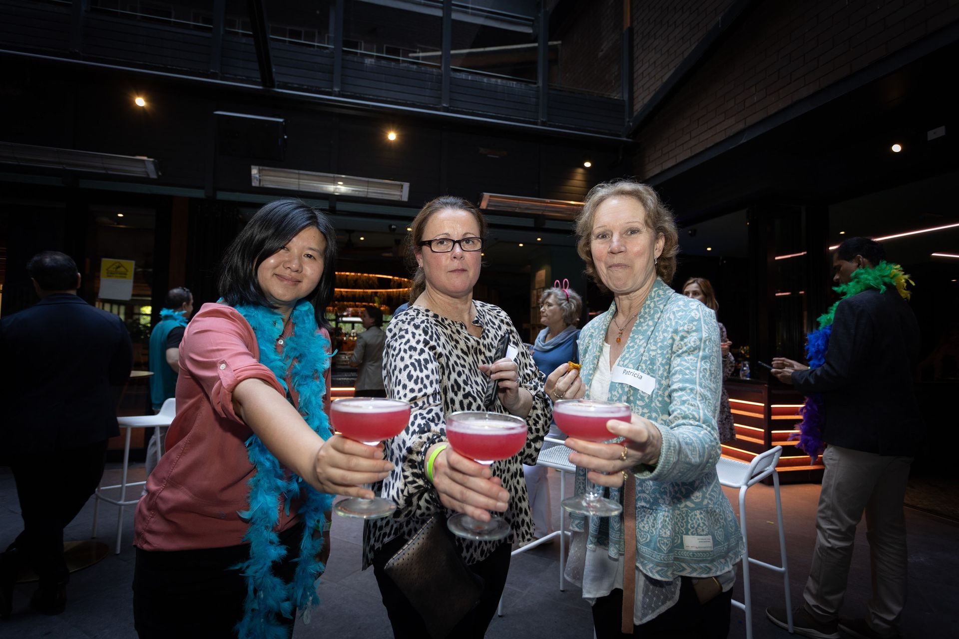 Three people at an indoor event hold up pink cocktails for a toast, smiling towards the camera.