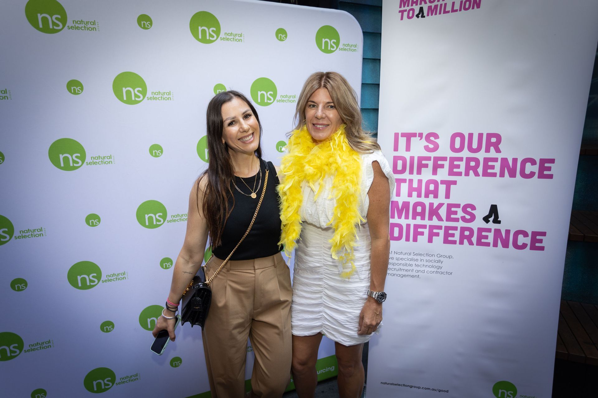 Two smiling people stand before a branded step-and-repeat banner. The person on the right wears a bright yellow boa.