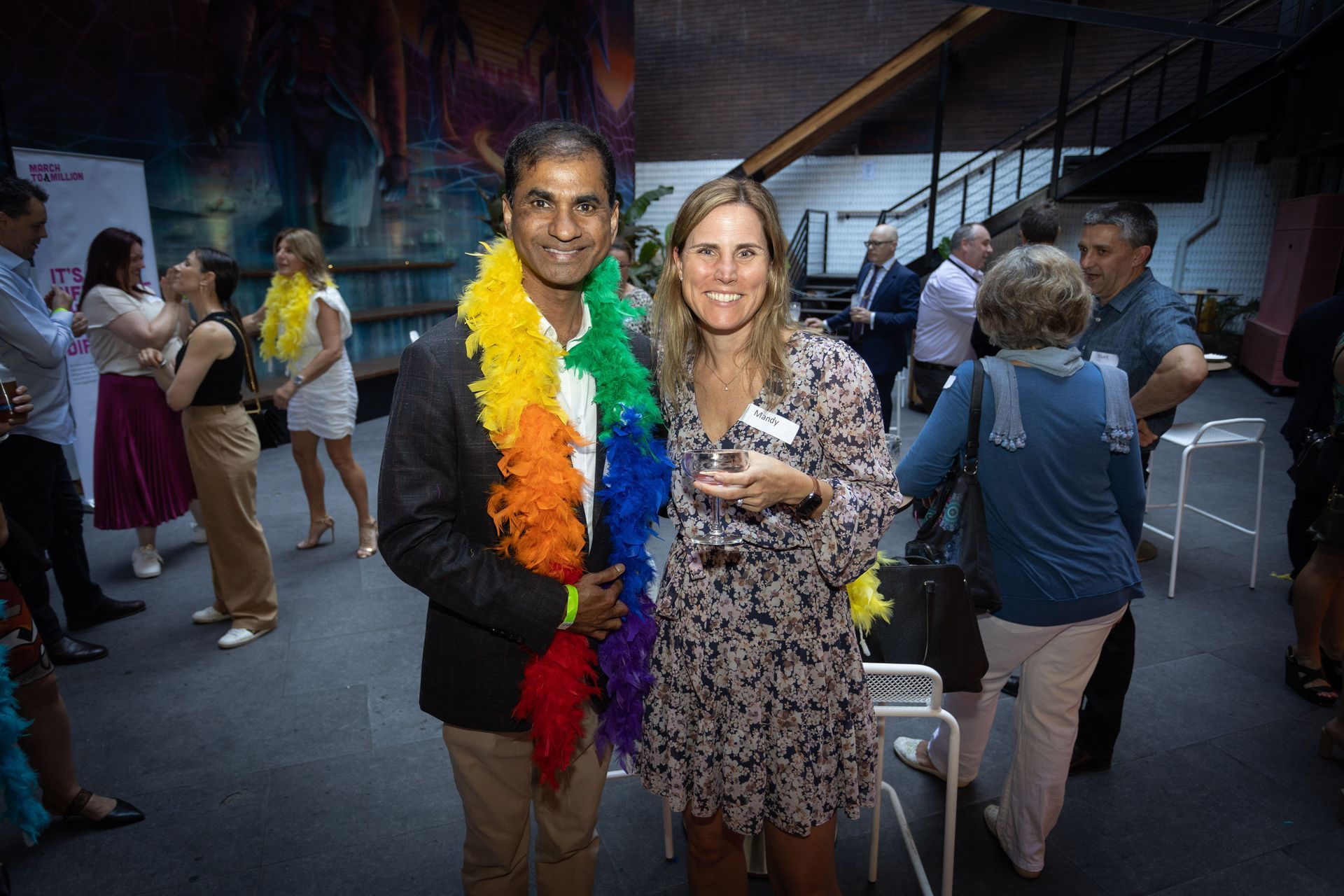 A person wearing a rainbow feather boa stands next to a person in a floral dress at an indoor event with other attendees.