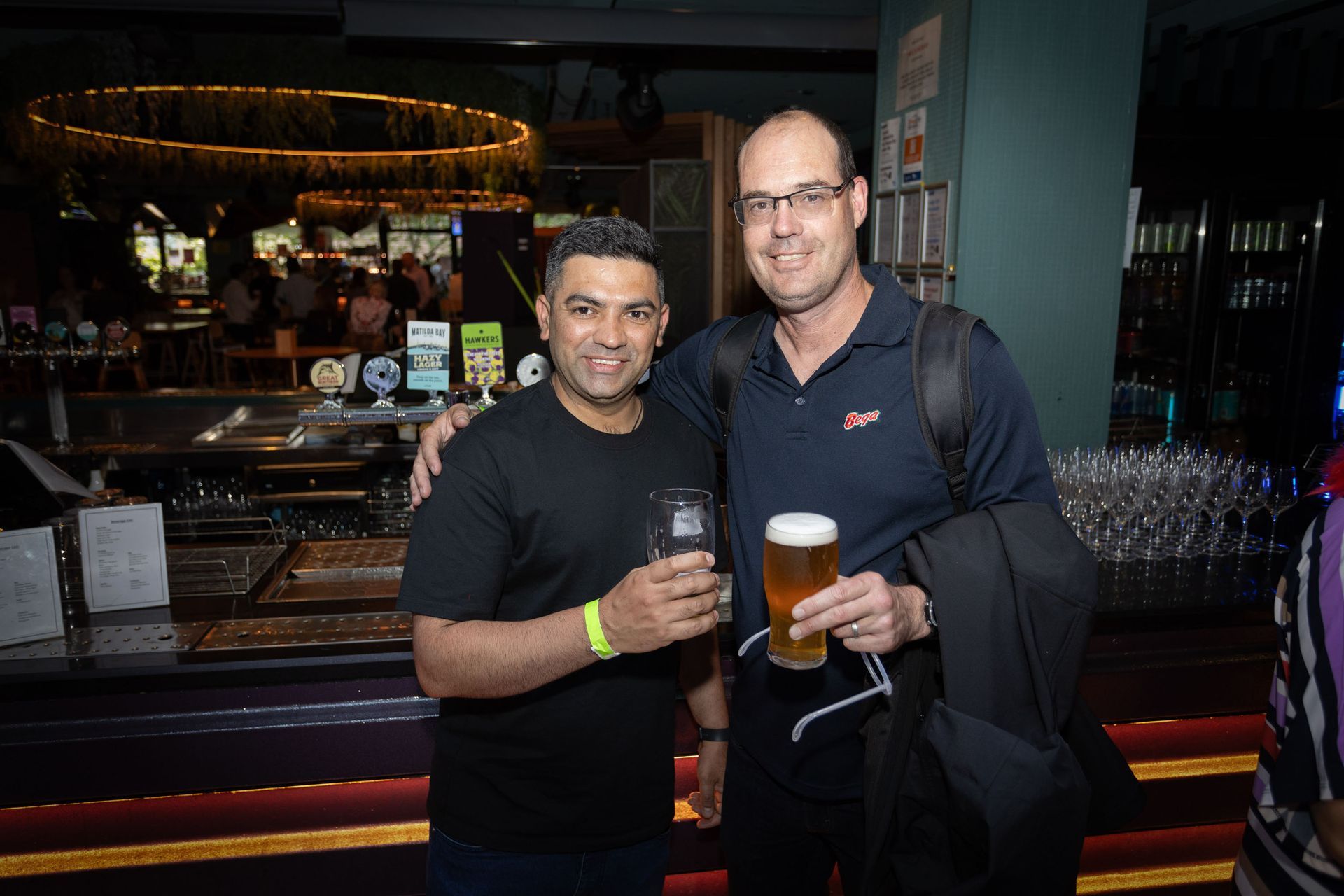Two men standing at a bar, both holding beer glasses, smiling, and looking toward the camera.