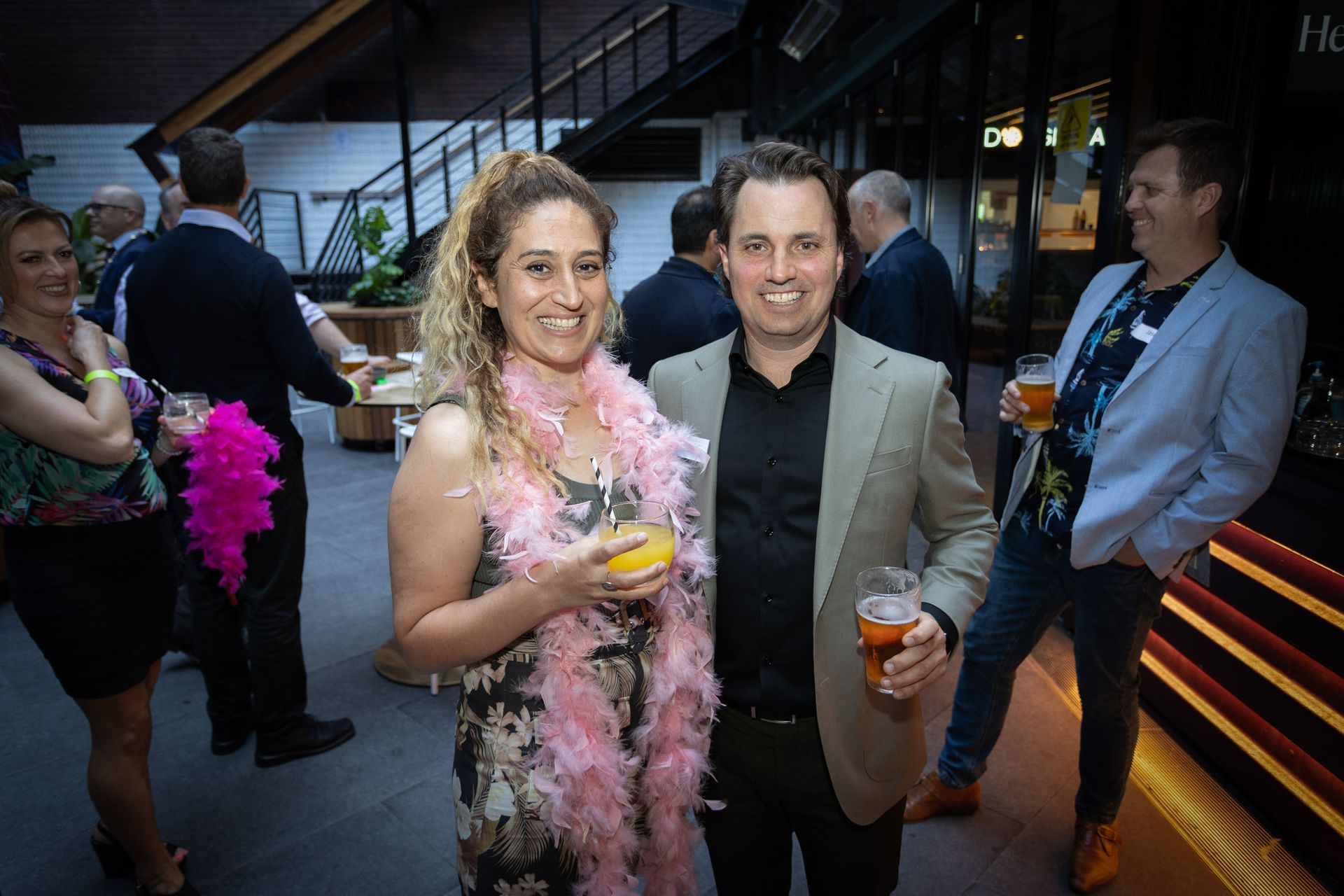 A woman in a pink feather boa and a man in a blazer pose with drinks at an indoor social event with other guests.