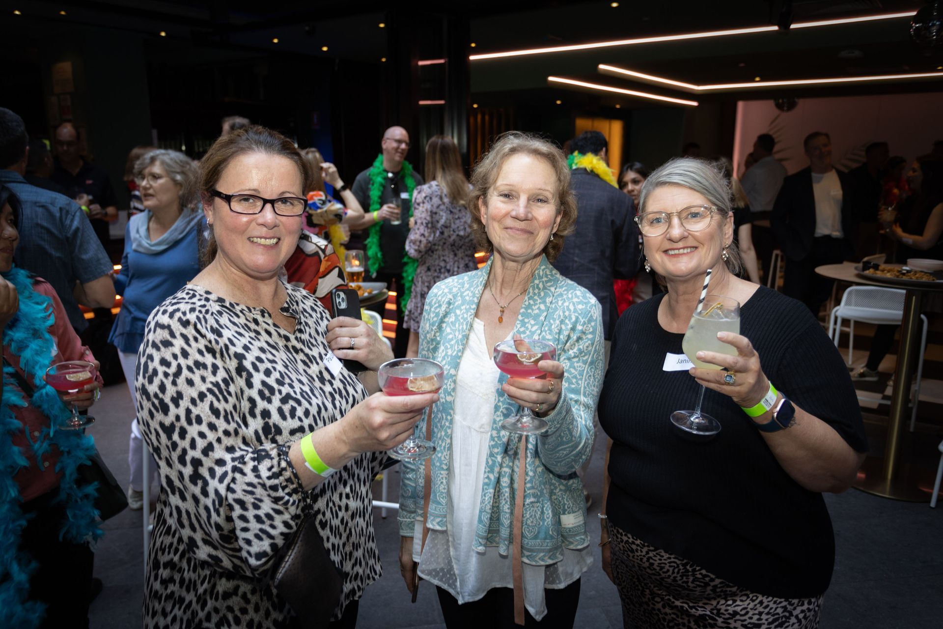 Three people smiling and holding cocktail glasses at a brightly lit indoor party with other guests in the background.