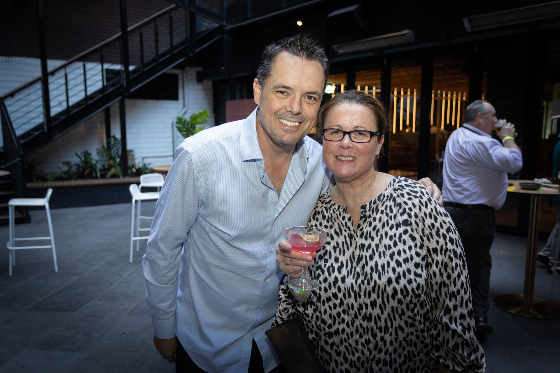 A smiling man and woman pose together with a cocktail in an outdoor bar area, with a person in the background.