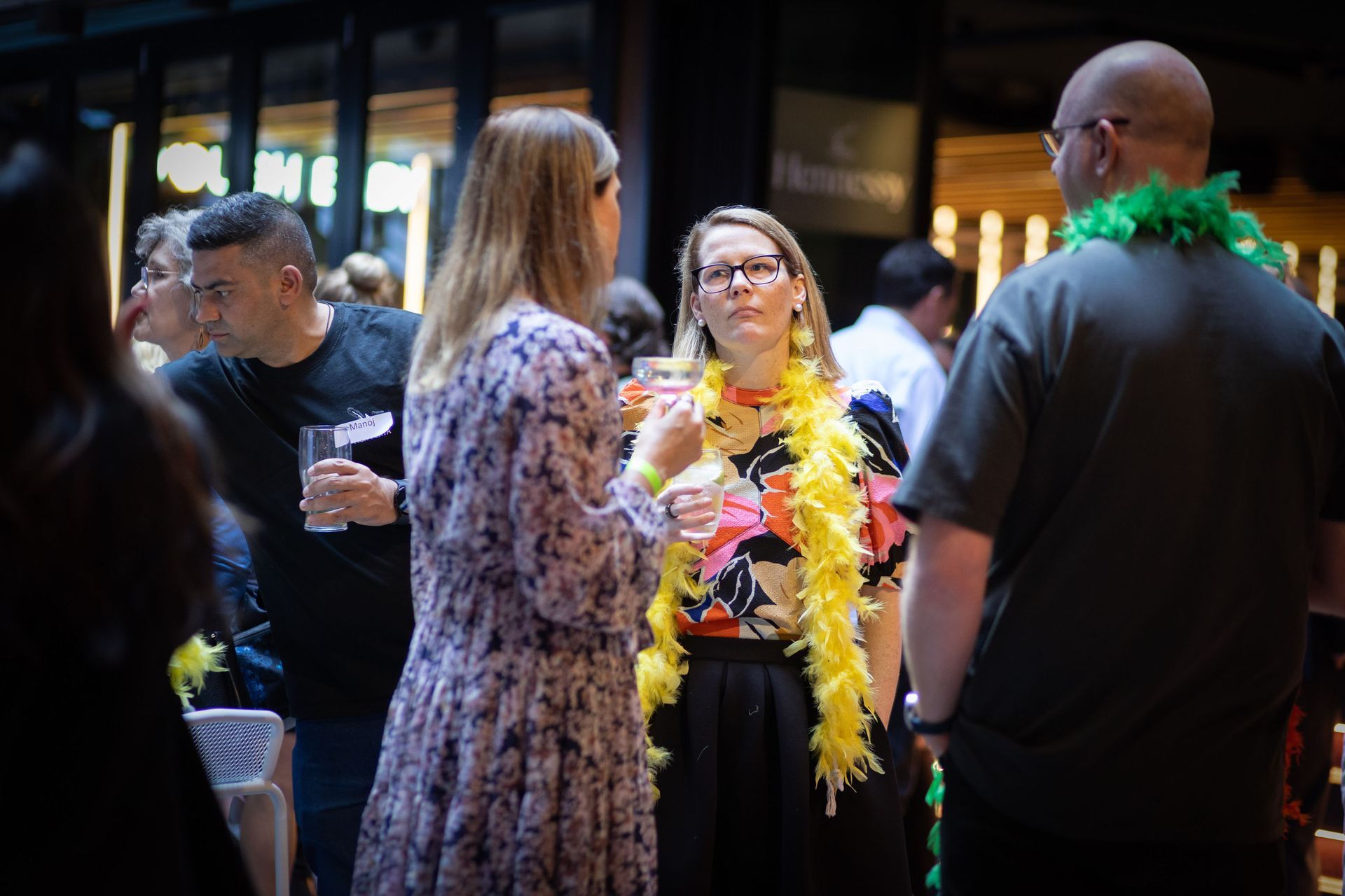 People in casual, festive attire with feather boas chat while holding drinks in a dimly lit, social event space.