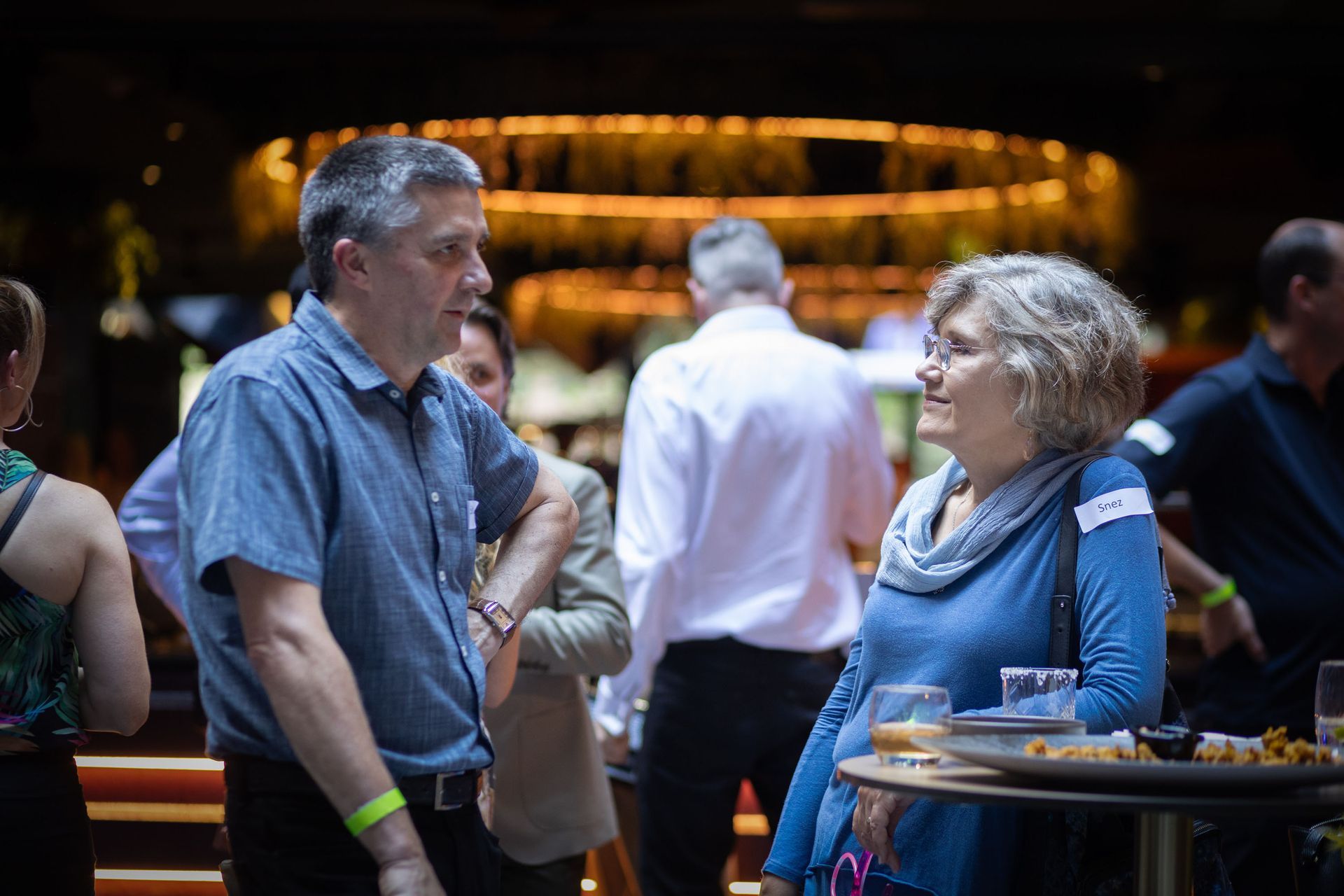 A man and a woman in blue shirts engage in a conversation at a social gathering with others standing nearby.