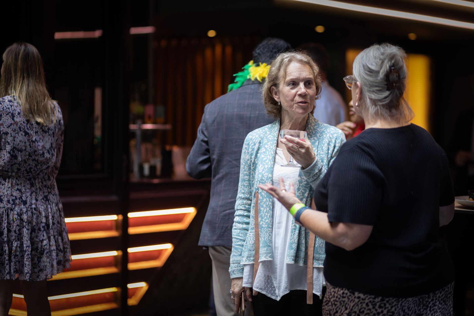 Three people converse at an indoor evening event with a bar in the background.