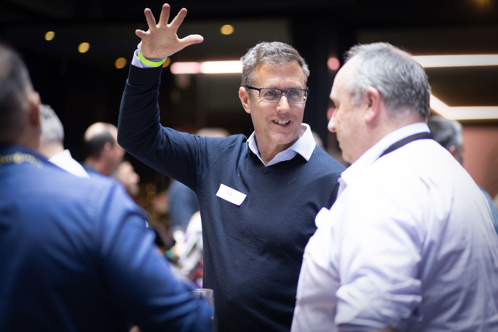A person in a dark sweater gestures while talking to others in a dimly lit, professional networking setting.