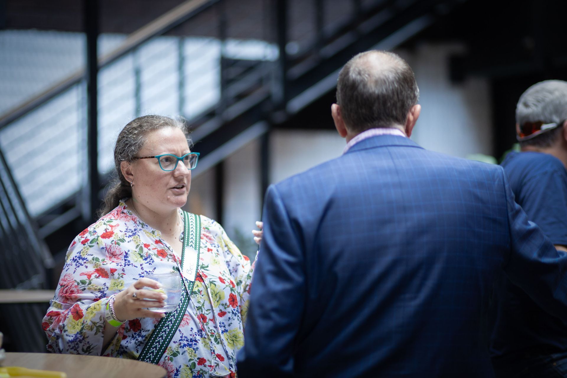 A person in a floral shirt speaks to another in a blue suit at an indoor event with a staircase in the background.