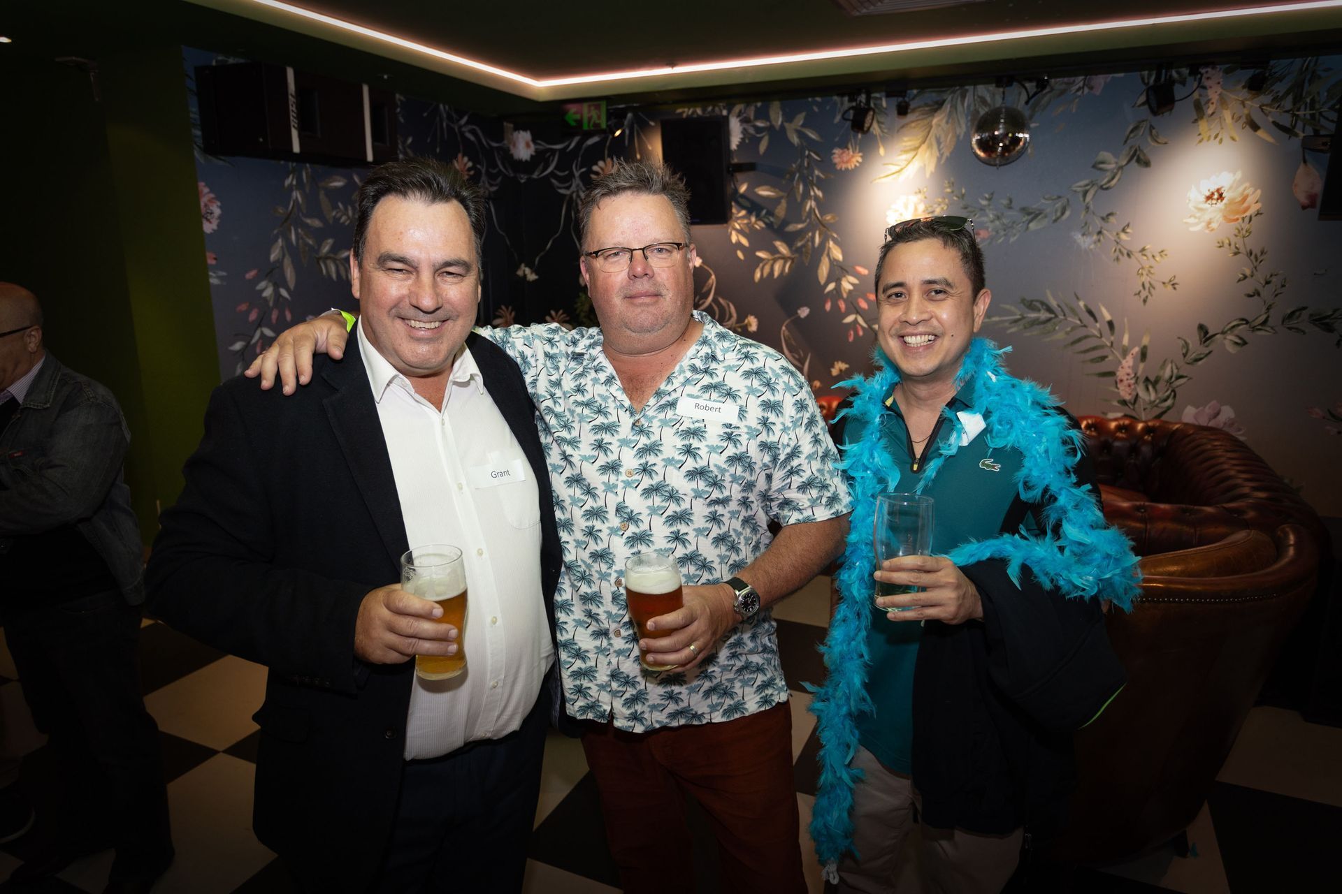 Three people smiling while holding glasses in a decorated lounge with a floral wall and checkered floor.