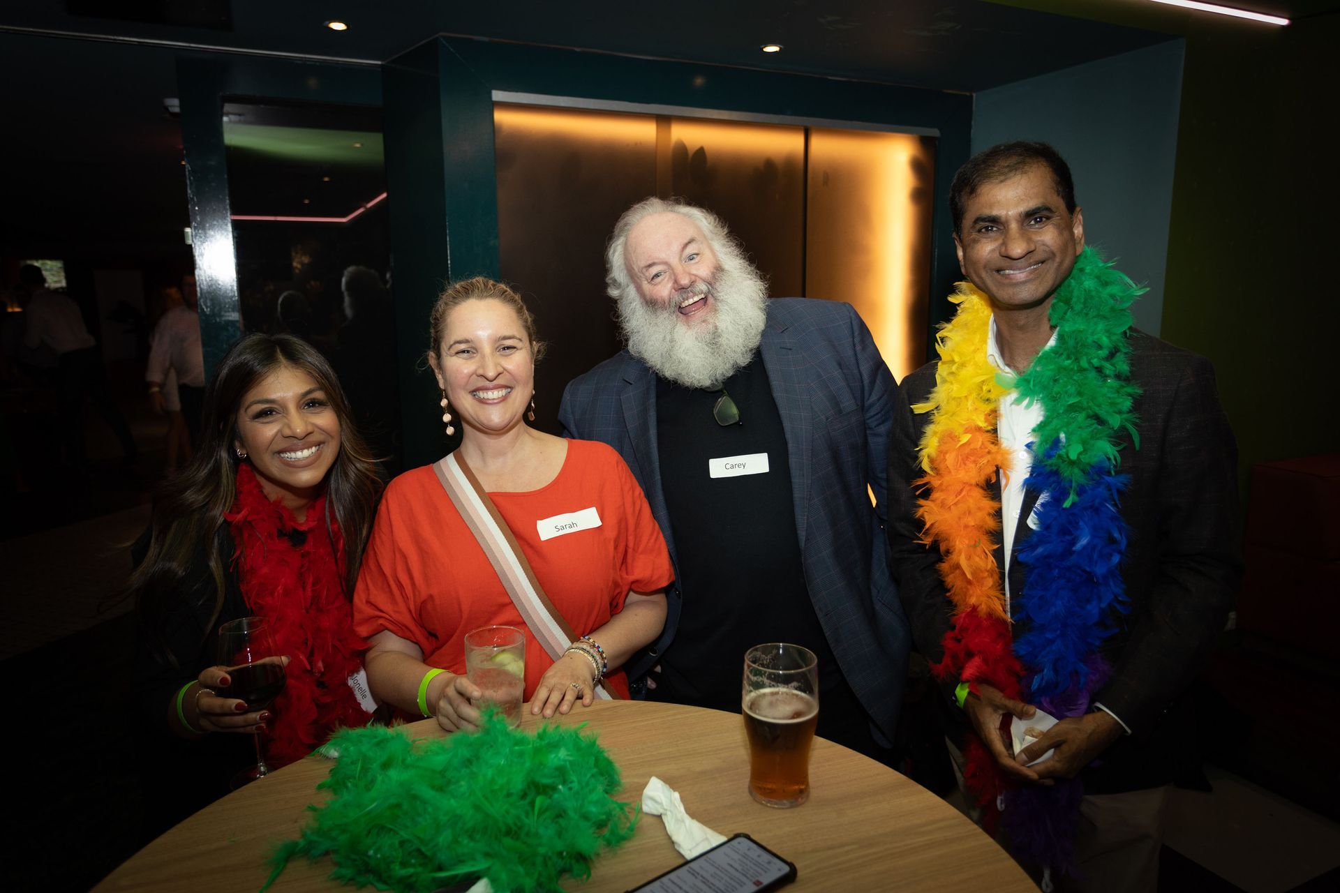 Four people smile and pose around a circular table in a dimly lit room, with one person wearing a rainbow feather boa.