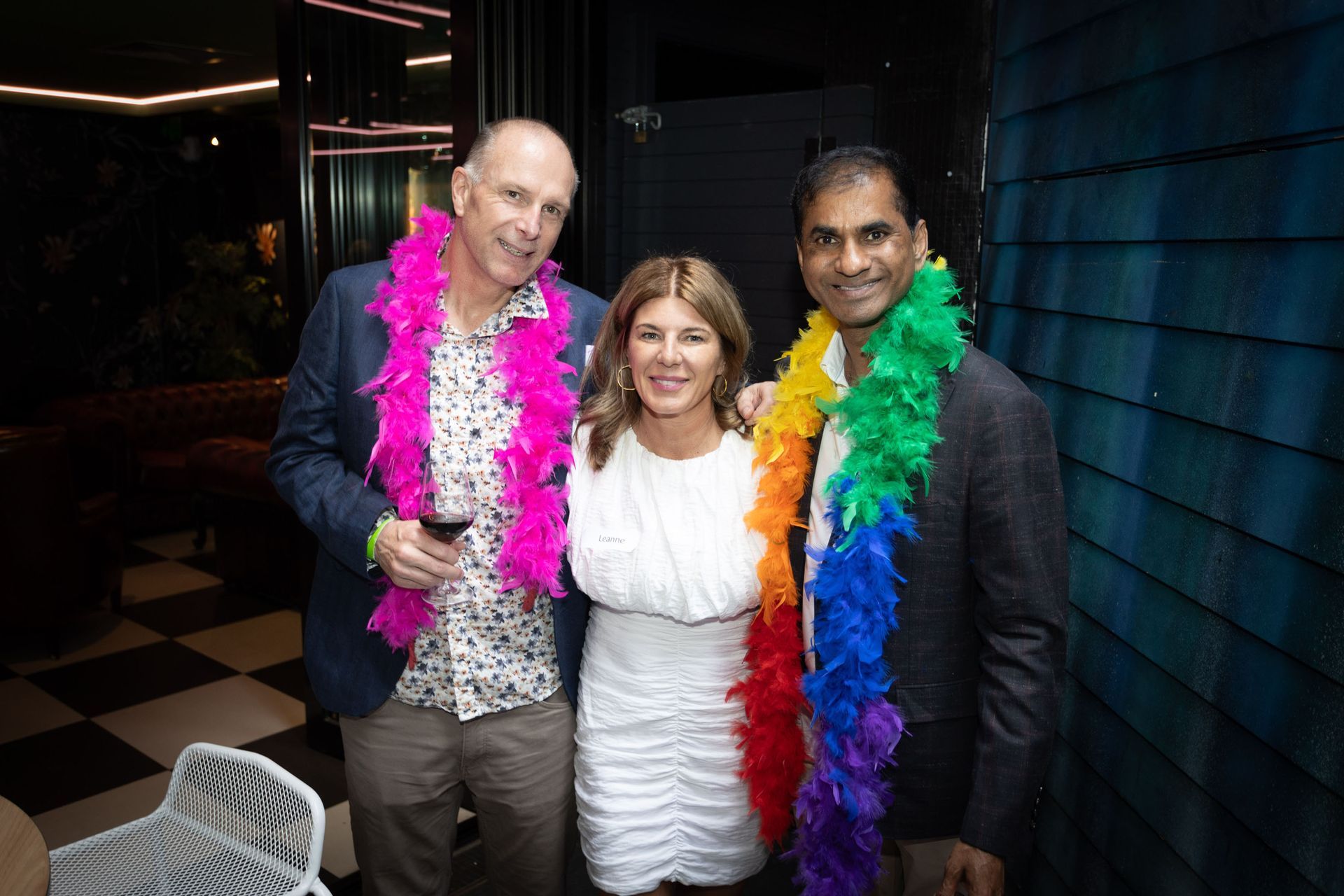 Three people pose together, smiling, wearing colorful feather boas inside a venue with checkered floors.