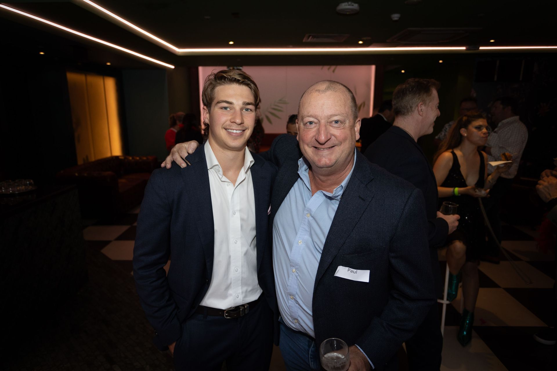 Two men in semi-formal business attire smile while standing close together in a dim, modern event space.
