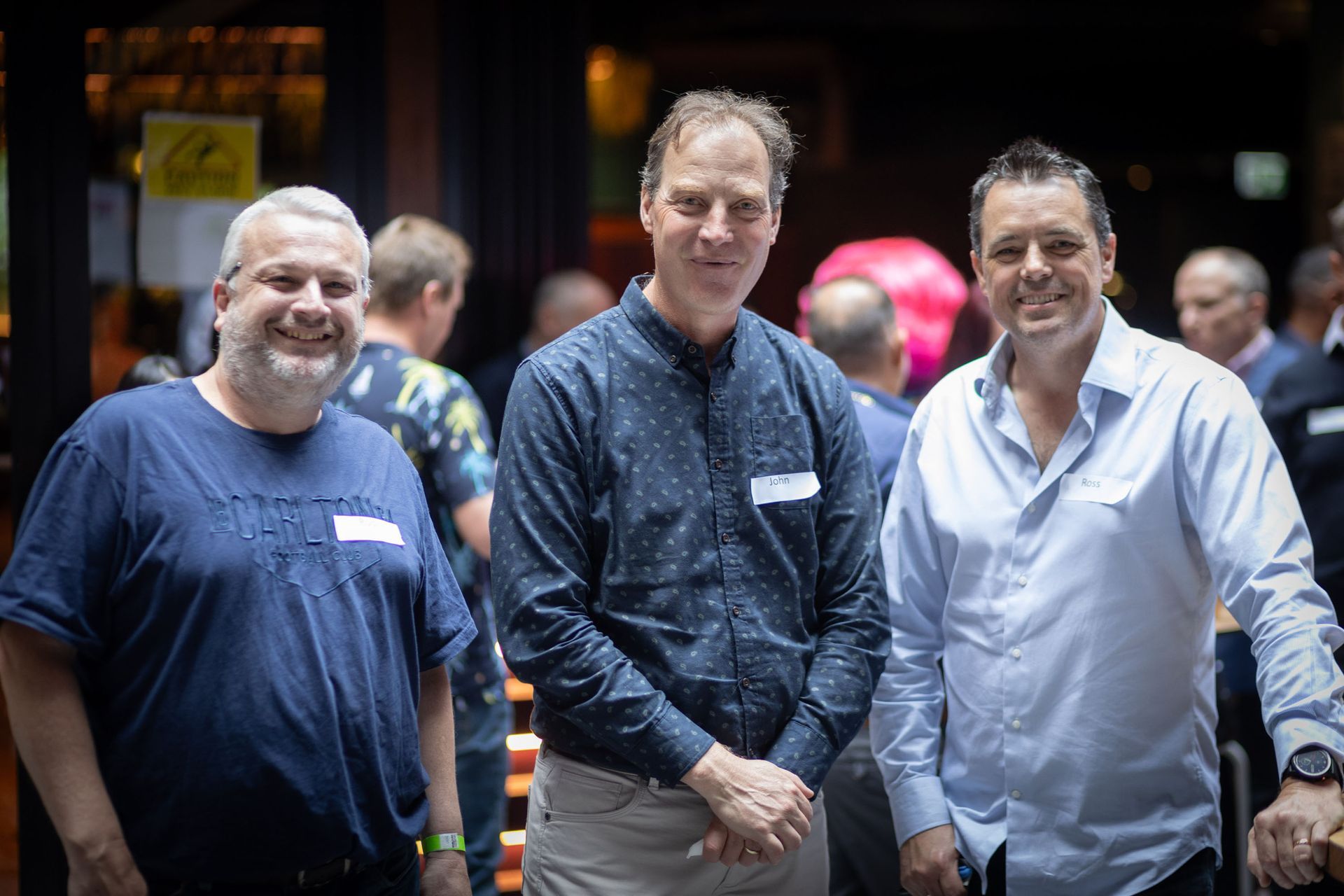 Three men smiling at a social gathering, standing side-by-side in a softly lit, indoor venue.