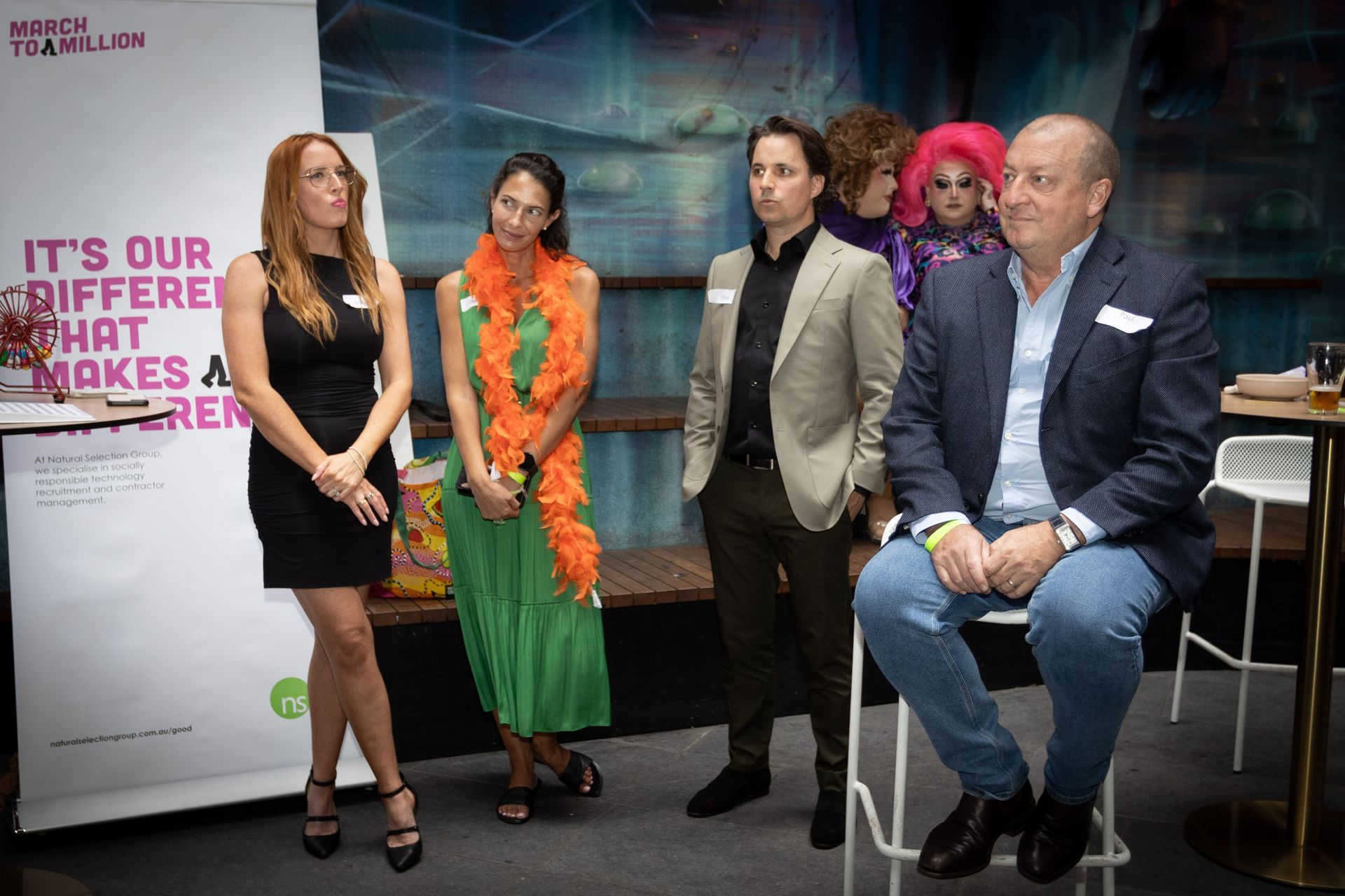 A group of four people stand on a stage next to a sign, with one man sitting on a stool.